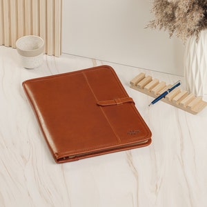 May include: A brown leather portfolio with a strap and a metal zipper. The portfolio is lying on a white surface with a wooden pen holder and a blue pen.
