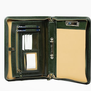 May include: A green leather zippered portfolio with a built-in clipboard, pen holders, and card slots. The portfolio is open and the interior is lined with a beige fabric.