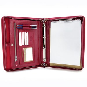 May include: A red leather zippered binder with a ring mechanism, a notepad, and pen holders. The binder is open and the inside is lined with a beige fabric.