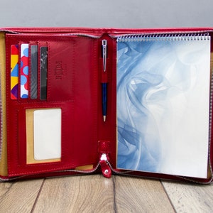 Può includere: Un portafoglio in pelle rossa con cerniera con un blocco note, una penna e slot per carte. Il blocco note presenta un design astratto blu e bianco.