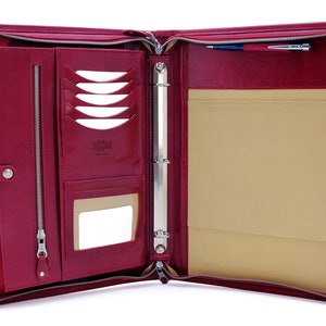 May include: A burgundy leather zippered binder with a ring mechanism, a pen holder, and multiple pockets. The binder is open and the interior is lined with a beige fabric.