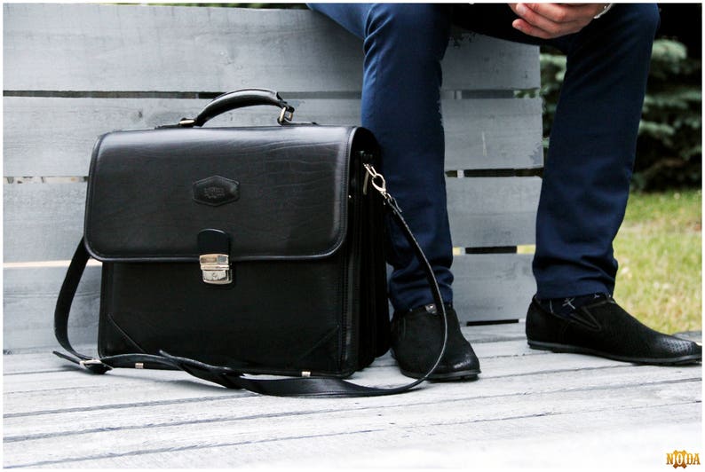 May include: A black leather briefcase with a silver clasp and a long strap. The briefcase is sitting on a wooden surface.
