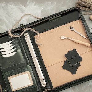 May include: A dark green leather binder with a zipper closure and multiple pockets. The binder is open and has a beige interior with a set of leather working tools, including a stitching awl, a stitching groover, a pricking iron, and a leather punch. The tools are laid out on a beige felt surface.