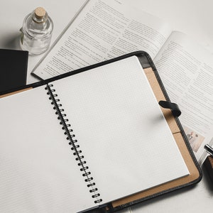 May include: An open spiral bound notebook with a black leather cover, lying on a white surface with open books and a glass bottle. The notebook has a grid pattern on the pages.