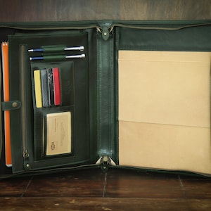 May include: A dark green leather portfolio with a zipper closure. The portfolio is open and has a notepad, pens, and business cards inside.