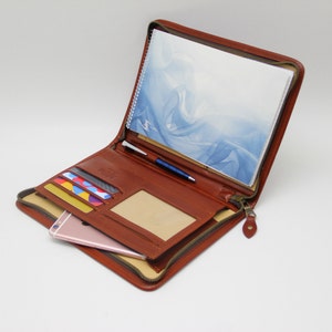 May include: A brown leather zippered portfolio with a notepad, pen, and credit cards inside. The portfolio has a zippered closure and a flap with a clear plastic window.