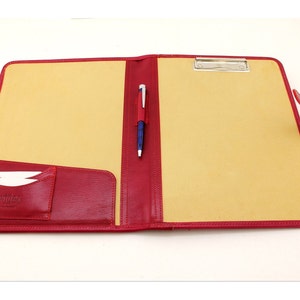 May include: Red leather folder with a yellow interior, a pen holder, and a small pocket on the front. The folder has a metal clip on the top.
