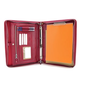 May include: A red leather zippered binder with a built-in notepad and pen holders. The binder is open and has a blank orange sheet of paper inside.
