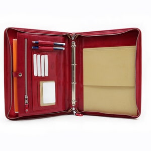 Puede incluir: Carpeta de cuero rojo con cremallera y mecanismo de anillas doradas. La carpeta está abierta y tiene un interior beige con bolsillos y un portaplumas. Hay dos bolígrafos en el portaplumas.