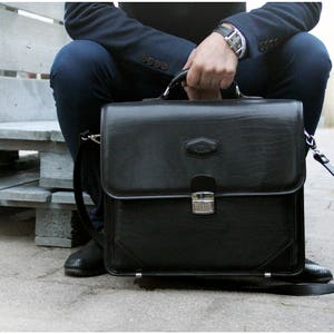 May include: A black leather briefcase with a silver clasp and a long strap. The briefcase is sitting on a wooden bench.