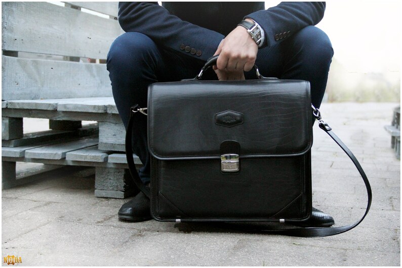 May include: A black leather briefcase with a silver clasp and a long strap. The briefcase is sitting on a wooden bench.