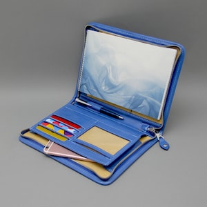 May include: A blue leather zippered travel wallet with multiple card slots, a pen holder, a phone pocket, and a clear ID window. The wallet is open and has a notebook inside.