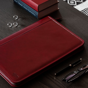 May include: A red leather zippered portfolio with a zipper pull that says "TUMI" on a dark wood surface.