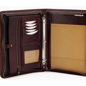 May include: A brown leather zippered binder with a ring mechanism, multiple pockets, and a pen holder. The binder is open and the interior is lined with a beige fabric.