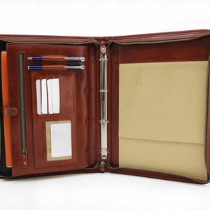 May include: A brown leather zippered binder with a ring mechanism for holding papers. The binder has a zippered pocket on the front and a clear plastic card holder on the inside.