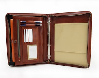 May include: A brown leather zippered binder with a ring mechanism for holding papers. The binder has a zippered pocket on the front and a clear plastic card holder on the inside.