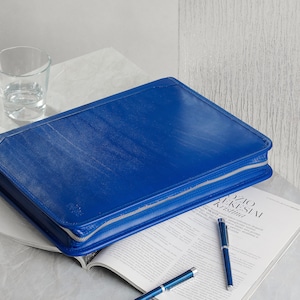 May include: A blue leather zippered portfolio on top of a magazine with two silver pens.