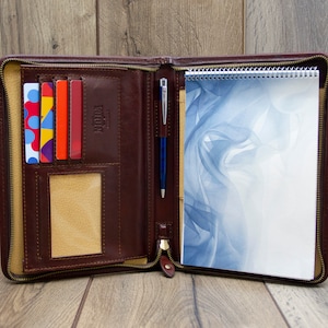 May include: A brown leather zippered portfolio with a notepad, pen, and card slots. The notepad has a blue and white abstract design.