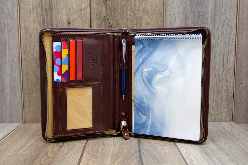 May include: A brown leather zippered portfolio with a notepad, pen, and card slots. The notepad has a blue and white abstract design.