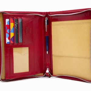May include: Red leather zippered portfolio with a pen holder, card slots, and a clear ID window. The interior is lined with a beige fabric.