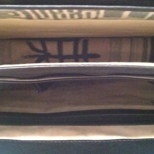 May include: Black leather bag with a zippered closure and a fabric lining. The bag is open and the lining is visible. The fabric lining has a pattern of black lines.