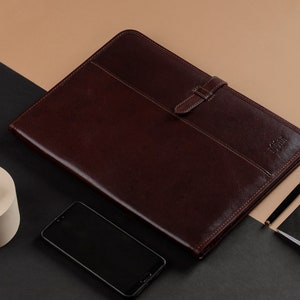 Peut inclure: Un porte-documents en cuir marron avec une fermeture à boucle. Un smartphone noir, un étui de recharge sans fil blanc, un stylo noir et un carnet noir avec le texte "FIELD NOTES" sont sur une surface noire et marron.