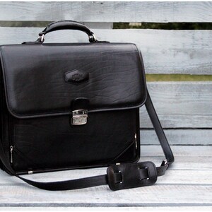 May include: A black leather briefcase with a silver clasp and a long adjustable strap. The briefcase has a textured surface and a small silver logo on the front.
