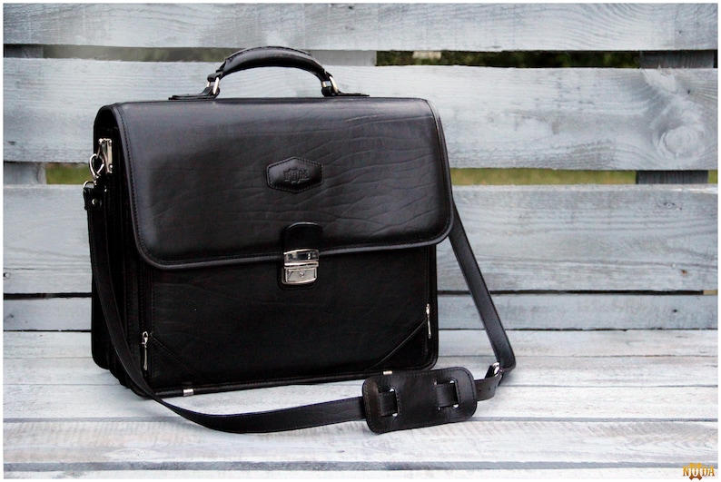 May include: A black leather briefcase with a silver clasp and a long adjustable strap. The briefcase has a textured surface and a small silver logo on the front.
