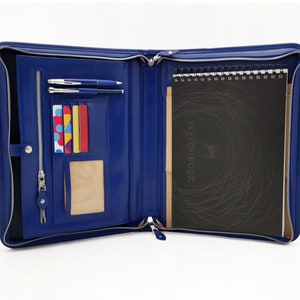 May include: A blue leather zippered portfolio with a notepad, pens, and card slots. The notepad has the word "SKETCHBOOK" printed on the cover.