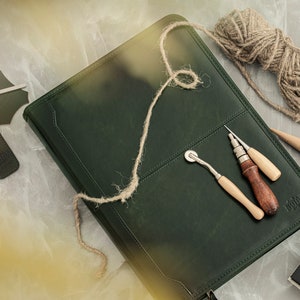 May include: A dark green leather journal with a brown leather strap and a metal clasp. The journal is surrounded by tools for leatherworking, including a stitching awl, a stitching groover, a leather punch, and a ball of twine.