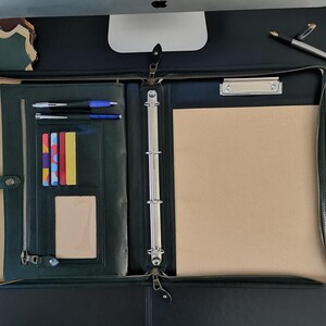 May include: A dark green leather zippered binder with a silver ring binder mechanism. The binder is open and has a beige notepad, a clear plastic card holder with colorful cards, and two black pens in the front pocket.