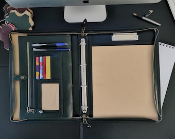 May include: A dark green leather zippered binder with a silver ring binder mechanism. The binder is open and has a beige notepad, a clear plastic card holder with colourful cards, and two black pens in the front pocket.