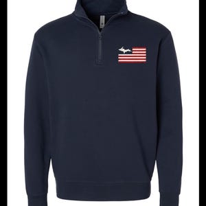 Peut inclure: Un sweat-shirt bleu marine à fermeture éclair quart de col avec un petit motif brodé sur la poitrine gauche. Le motif représente un drapeau américain stylisé où le champ bleu est remplacé par une silhouette blanche de la péninsule supérieure du Michigan.