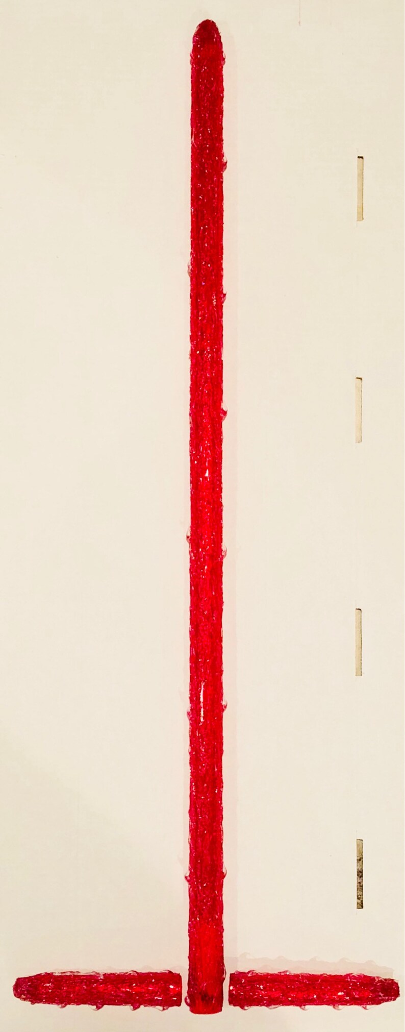 Op de afbeelding: Een rood, glinsterend, plastic, snoepstokvormig ornament met een lang, dun, verticaal gedeelte en drie kortere, horizontale gedeelten.