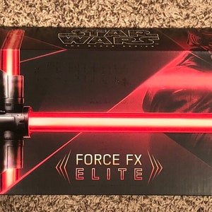 Op de afbeelding: Een zwarte doos met een rood lichtzwaard en de tekst "Supreme Leader Kylo Ren Force FX Elite Lightsaber" op de voorkant. De doos heeft ook het Star Wars logo en de tekst "Force FX Elite".