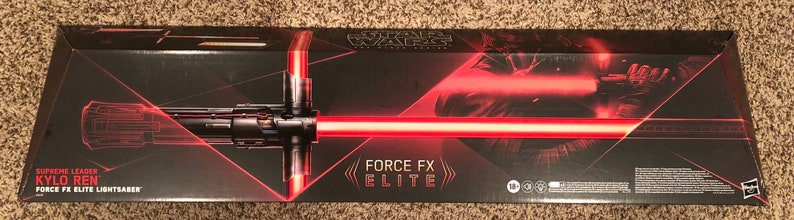 Op de afbeelding: Een zwarte doos met een rood lichtzwaard en de tekst "Supreme Leader Kylo Ren Force FX Elite Lightsaber" op de voorkant. De doos heeft ook het Star Wars logo en de tekst "Force FX Elite".