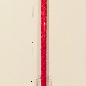 Op de afbeelding: Een rood lichtzwaard met een zwarte handgreep en een transparante plastic standaard. Het lichtzwaard is gemonteerd op de standaard met de kling naar boven gericht.