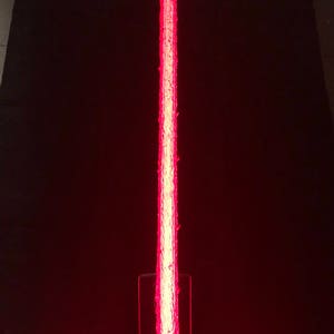 Op de afbeelding: Een rood lichtzwaard met een getextureerd blad en een zwarte handgreep. Het lichtzwaard is aan en het blad gloeit.