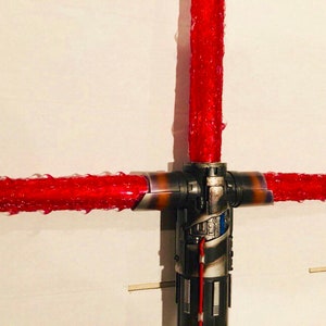 Op de afbeelding: Een rood lichtzwaard met een zwart en zilveren handvat. Het lichtzwaard bevindt zich in een kruisbeschermingsconfiguratie, met twee bladen die zich uitstrekken vanaf de zijkanten van het handvat.