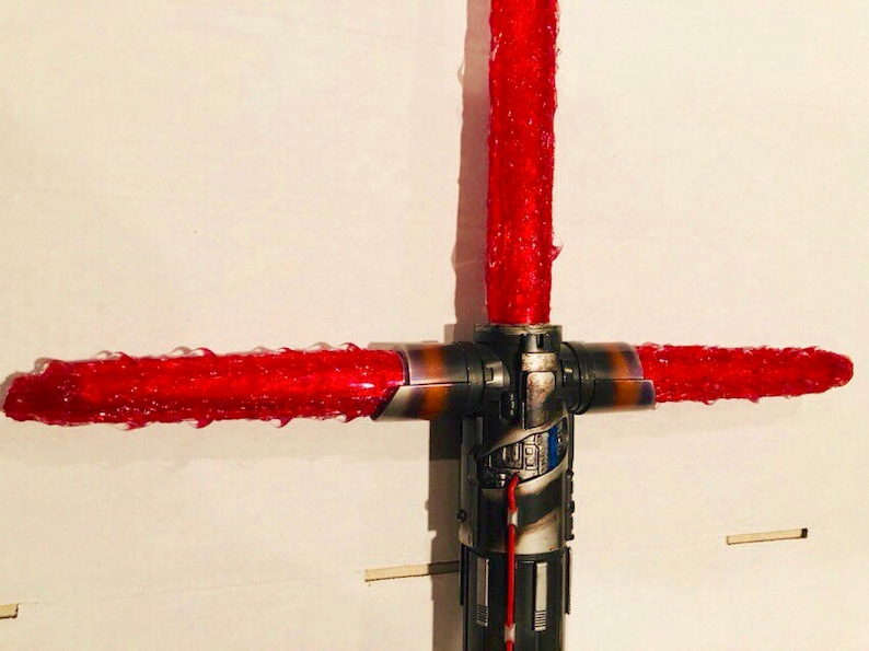 Op de afbeelding: Een rood lichtzwaard met een zwart en zilveren handvat. Het lichtzwaard bevindt zich in een kruisbeschermingsconfiguratie, met twee bladen die zich uitstrekken vanaf de zijkanten van het handvat.