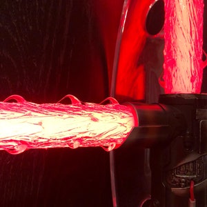 Op de afbeelding: Een rode lightsaber-prop met een transparante acrylstandaard. De lightsaber is aan en de blade is gemaakt van een doorschijnend materiaal.