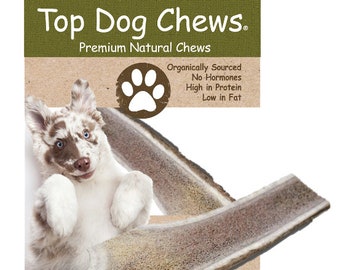 May include: A brown and white dog looks up at a long, light brown antler dog chew. The chew is on a brown background with the text "Top Dog Chews. Premium Natural Chews" and a paw print logo. The text below the logo reads "Organically Sourced No-Hormones High in Protein Low in Fat."
