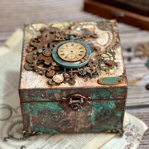 Puede incluir: Una caja decorativa cuadrada con acabado desgastado en color verde azulado y marr&oacute;n, con un cierre met&aacute;lico. La tapa est&aacute; adornada con engranajes met&aacute;licos, una esfera de reloj circular y una peque&ntilde;a placa ovalada con texto grabado.