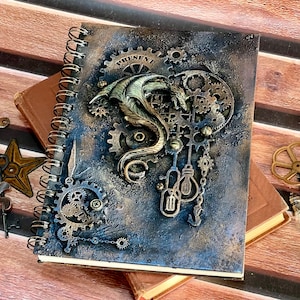 May include: A spiral-bound notebook with a textured, dark blue and bronze cover featuring a relief of a dragon, various mechanical gears, and two light bulb shapes. The word PRESENT is printed at the top. The cover has a metallic, weathered finish.