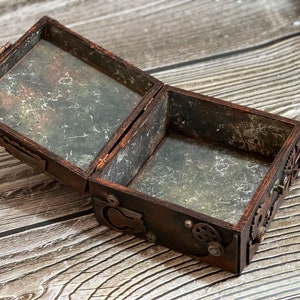 Puede incluir: Una caja rectangular de madera con la tapa abierta, con un acabado marr&oacute;n oscuro y adornos de engranajes met&aacute;licos en el exterior. Las superficies interiores tienen una textura desgastada de color gris y negro, con una bisagra en la parte trasera.