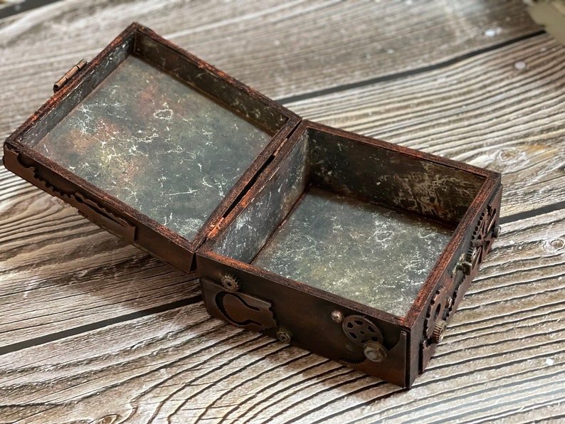 Puede incluir: Una caja rectangular de madera con la tapa abierta, con un acabado marr&oacute;n oscuro y adornos de engranajes met&aacute;licos en el exterior. Las superficies interiores tienen una textura desgastada de color gris y negro, con una bisagra en la parte trasera.