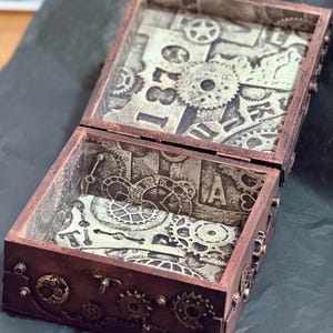 Puede incluir: Una caja rectangular de madera con tapa abatible, con un acabado en tono cobre oscuro y adornos de engranajes en el exterior. El interior est&aacute; forrado con papel que muestra un patr&oacute;n monocrom&aacute;tico de engranajes mec&aacute;nicos y el n&uacute;mero 1870.