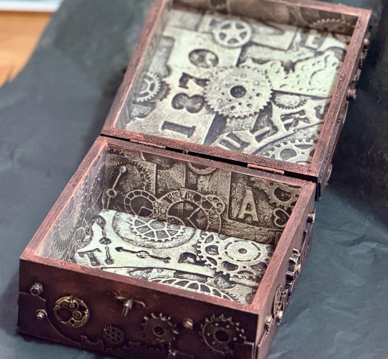 Puede incluir: Una caja rectangular de madera con tapa abatible, con un acabado en tono cobre oscuro y adornos de engranajes en el exterior. El interior est&aacute; forrado con papel que muestra un patr&oacute;n monocrom&aacute;tico de engranajes mec&aacute;nicos y el n&uacute;mero 1870.