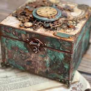 Puede incluir: Una caja decorativa cuadrada con un acabado desgastado en color verde azulado y marr&oacute;n, con un cierre de metal, esquinas ornamentadas y una tapa adornada con varios engranajes met&aacute;licos y una esfera de reloj circular con n&uacute;meros.