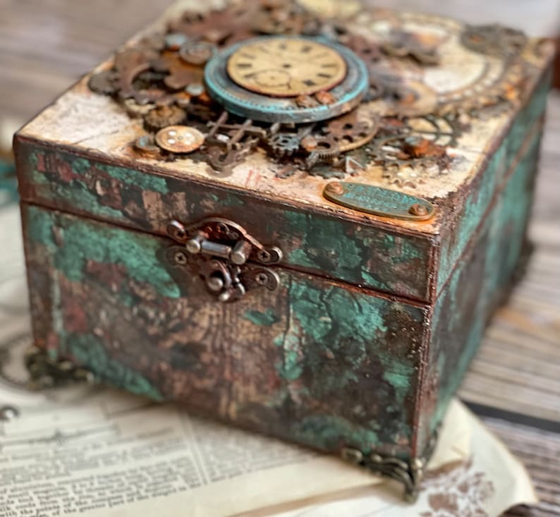 Puede incluir: Una caja decorativa cuadrada con un acabado desgastado en color verde azulado y marr&oacute;n, con un cierre de metal, esquinas ornamentadas y una tapa adornada con varios engranajes met&aacute;licos y una esfera de reloj circular con n&uacute;meros.